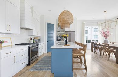Bright modern farmhouse kitchen with blue island, white shaker cabinets, stainless appliances, rattan pendant lights, wooden bar stools and open-plan dining with hardwood floors