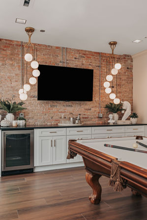 Modern home game room with wooden pool table, exposed brick accent wall, wall-mounted TV, globe pendant lights, white cabinets and mini-fridge on hardwood floors.