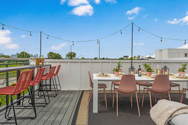 Urban rooftop deck with skyline views — white dining table set with pink chairs, red bar stools at the railing, string lights overhead and blue sky.