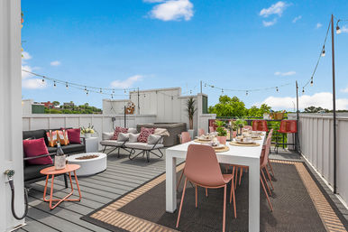 Urban rooftop deck with a white dining table set for eight and pink chairs, cozy lounge area with cushions and round fire pit, string lights and bar stools, overlooking treetops and city skyline under a bright blue sky.