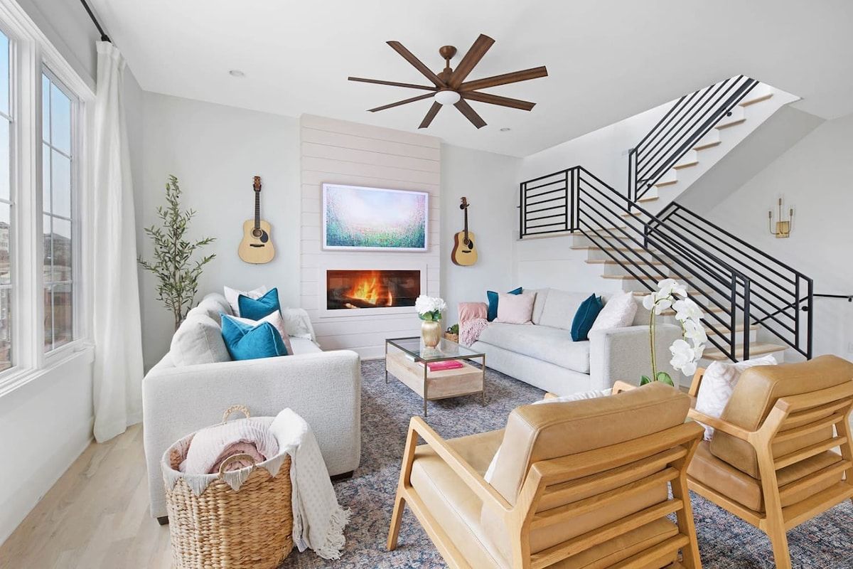 Sunlit modern living room with white sofas, teal pillows, leather lounge chairs, wall-mounted guitars, fireplace and TV, large window and black metal open staircase