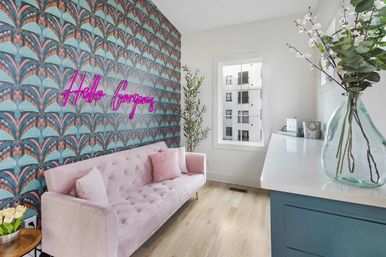 Bright modern apartment lounge with pink velvet tufted sofa, teal art-deco wallpaper and neon “Hello gorgeous” sign, light wood floors, large window and plants.