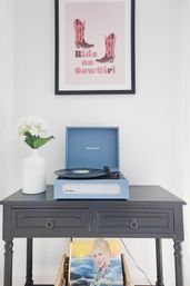 Vinyl turntable home decor: blue portable record player on a black console table with a white hydrangea vase, framed pink cowgirl print above, and a vinyl album stored below.