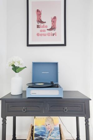 Vinyl turntable home decor: blue portable record player on a black console table with a white hydrangea vase, framed pink cowgirl print above, and a vinyl album stored below.