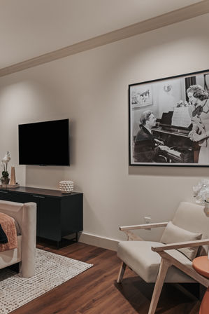 Cozy modern bedroom sitting area with wall-mounted TV, black media console, framed black-and-white piano print, beige mid-century armchair, textured rug and warm hardwood floors.