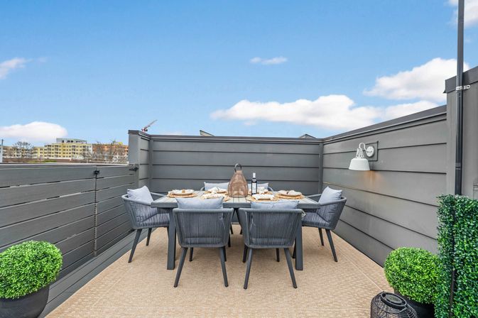 Cozy rooftop terrace with gray privacy panels and a modern six-seat outdoor dining set — woven cushioned chairs around a table staged with plates, wine bottle and lantern centerpiece, potted boxwood accents and distant urban skyline under a blue sky.