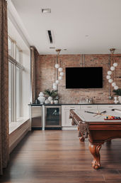 Sunlit industrial-style loft game room with a wooden pool table, exposed brick accent wall, wall-mounted TV, globe pendant lights, wet bar with mini-fridges and decorative vases, and large windows letting in natural light.