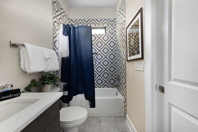 Modern small bathroom featuring a geometric black-and-white tile accent wall, navy-and-white shower curtain over a white bathtub, white countertop vanity with folded towels and a potted plant, toilet and framed wall art.