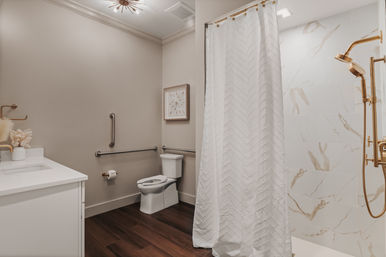 Modern accessible bathroom interior with white vanity, toilet with horizontal and vertical grab bars, walk-in shower behind a textured white curtain with marble-look tile and gold fixtures, dark wood-look flooring and neutral walls.