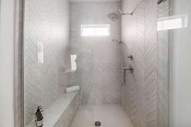 Glass-enclosed walk-in shower with light gray herringbone tile, built-in bench with rolled towel, chrome rainfall and handheld showerheads, white mosaic floor, and a small high window letting in natural light.
