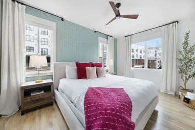 Bright modern city apartment bedroom with white upholstered bed, white bedding and magenta throw and pillows, teal patterned accent wall, wood nightstand, large windows with city views, ceiling fan and light hardwood floors.
