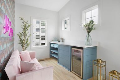 Sunny modern apartment lounge with pink velvet sofa, patterned accent wall and neon sign, tall potted plant, blue built-in cabinet with white countertop and under-counter beverage fridge, glass vase of greenery, hardwood floors and windows with city view.
