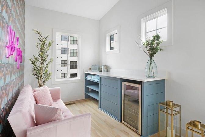 Sunny modern apartment lounge with pink velvet sofa, patterned accent wall and neon sign, tall potted plant, blue built-in cabinet with white countertop and under-counter beverage fridge, glass vase of greenery, hardwood floors and windows with city view.