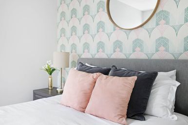 Bright modern bedroom with gray upholstered headboard, pastel art-deco wallpaper, round mirror, gold bedside lamp and pink and charcoal throw pillows