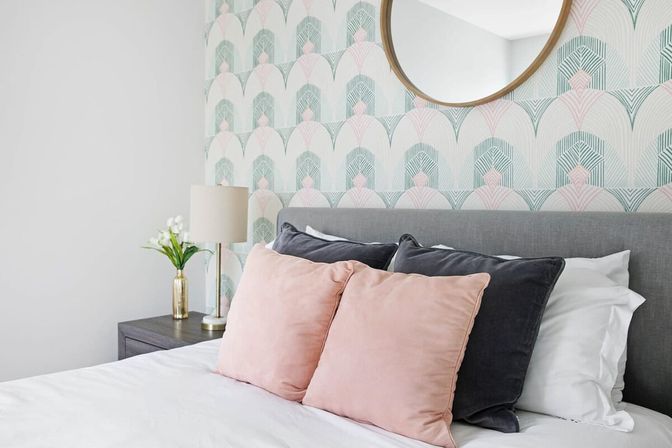 Bright modern bedroom with gray upholstered headboard, pastel art-deco wallpaper, round mirror, gold bedside lamp and pink and charcoal throw pillows