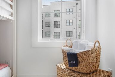 Cozy bright minimalist apartment corner with a rain-speckled window and natural woven basket of rolled white towels on a wicker trunk