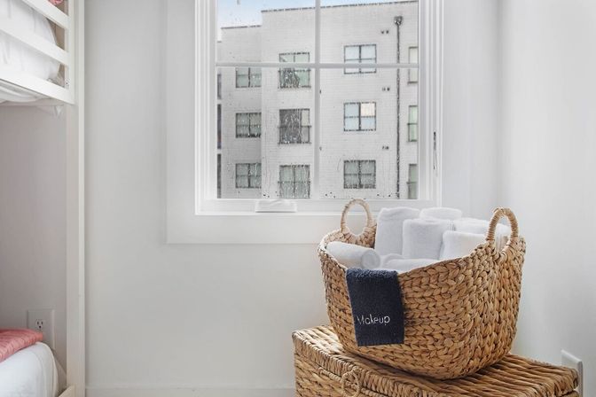 Cozy bright minimalist apartment corner with a rain-speckled window and natural woven basket of rolled white towels on a wicker trunk