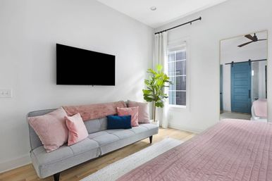 Bright modern bedroom with light-gray sofa dressed in pink and navy throw pillows, wall-mounted flat-screen TV, pink quilted bedspread, potted green plant by a sunlit window, and a large mirror reflecting a teal sliding barn door.