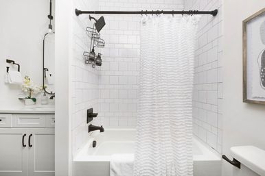 Crisp modern bathroom featuring a white subway-tile tub with a ruffled white shower curtain, matte black shower fixtures and caddy, and a white vanity with orchid and mirror.