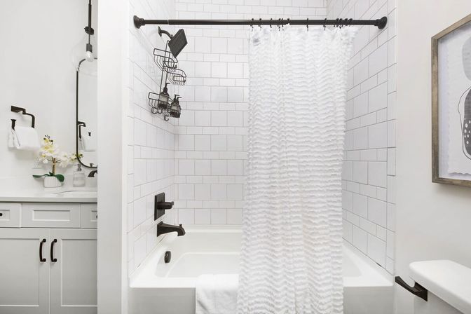 Crisp modern bathroom featuring a white subway-tile tub with a ruffled white shower curtain, matte black shower fixtures and caddy, and a white vanity with orchid and mirror.