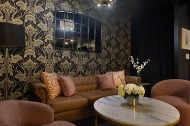 Cozy chic living room interior with gold floral wallpaper, tufted brown leather sofa, pink boucle armchairs, marble coffee table topped with white hydrangeas and a grid wall mirror.