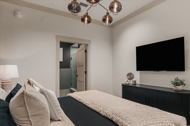 Cozy modern master bedroom with plush neutral bedding, textured throw, wall-mounted flat-screen TV, black media console, globe chandelier, and open ensuite glass shower.