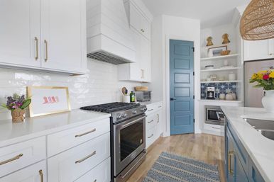 Bright modern coastal kitchen with white shaker cabinets and brass hardware, subway tile backsplash, stainless steel gas range, blue paneled door and island, open shelving with patterned tile, woven pendant and hardwood floors.