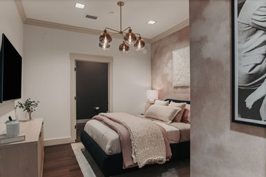 Cozy modern bedroom interior in blush and neutral tones with a layered queen bed, chunky knit throw, brass multi-globe pendant light, textured wall art, bedside lamp, TV on a dresser, and dark hardwood floors.