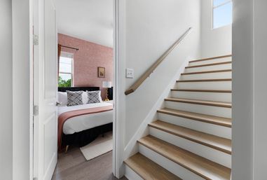 Modern home interior with wood-tread staircase next to a partially open door revealing a stylish bedroom with pink patterned accent wall, black upholstered headboard, geometric pillows and a brass bedside lamp.