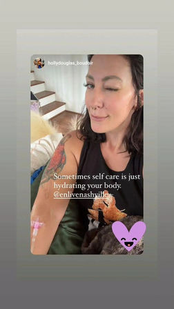 Winking woman with septum piercing and colorful arm tattoo relaxing on a couch with a visible hydration IV in her forearm, cozy living room setting with wooden stairs and overlaid caption about self-care and hydrating your body.