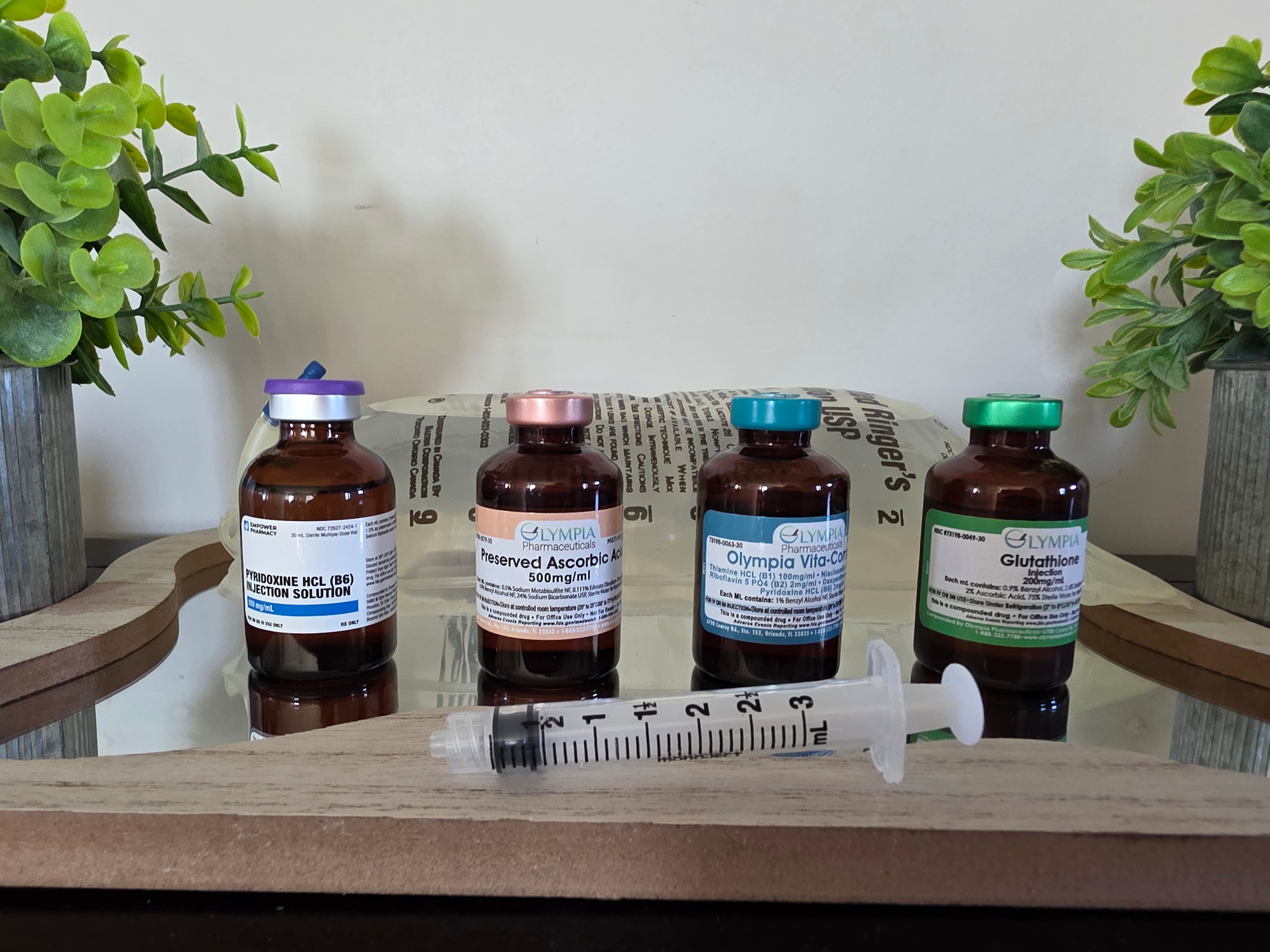 Amber glass vials with colorful caps and labels lined on a wooden tray with a 3ml syringe in front, flanked by potted green plants — IV vitamin/injection wellness setup.