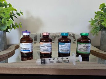 Amber glass vials with colorful caps and labels lined on a wooden tray with a 3ml syringe in front, flanked by potted green plants — IV vitamin/injection wellness setup.