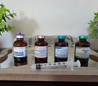 Amber glass vials with colorful caps and labels lined on a wooden tray with a 3ml syringe in front, flanked by potted green plants — IV vitamin/injection wellness setup.