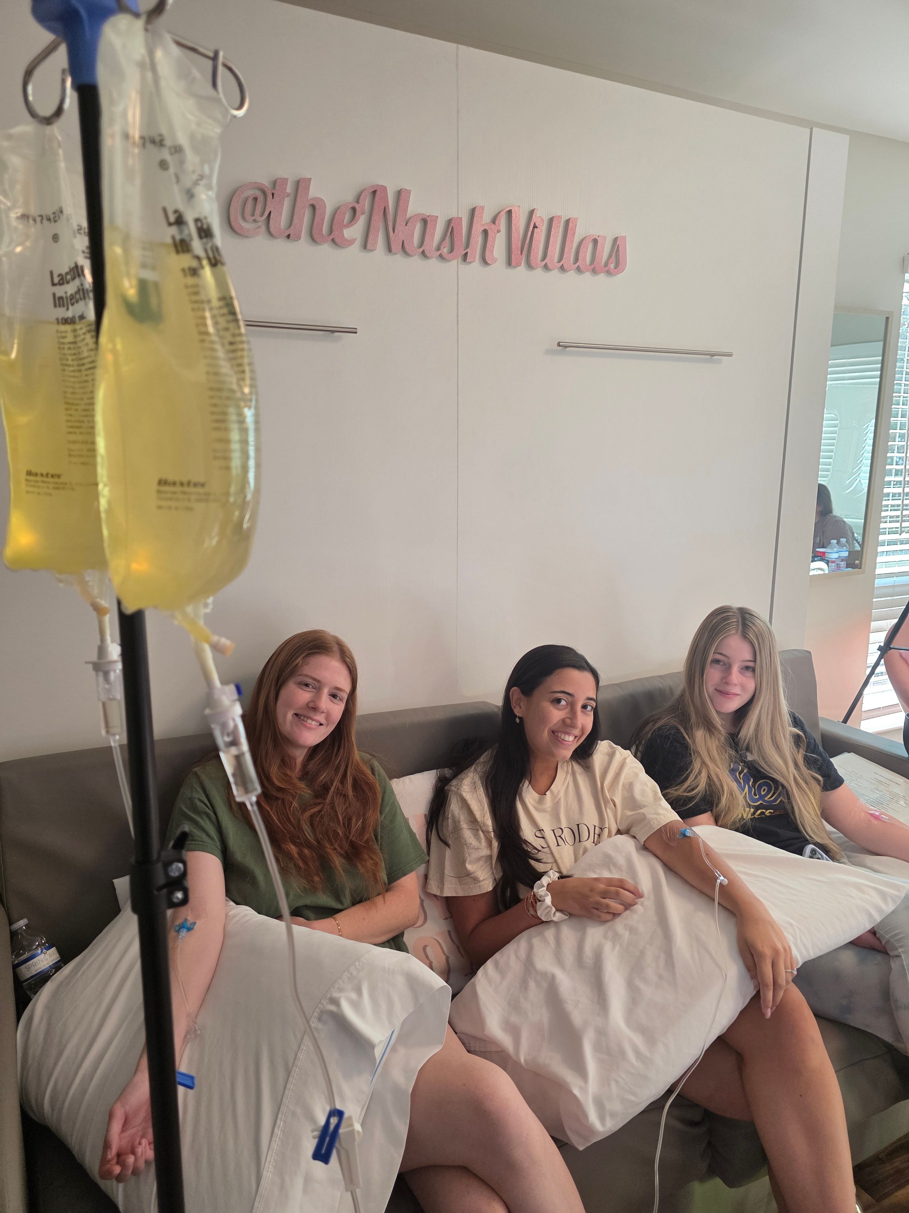 Three smiling women relax on a couch in a bright wellness lounge receiving IV vitamin drips, holding pillows while IV lines and yellow infusion bags hang in the foreground.