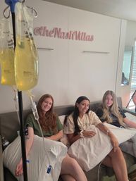 Three smiling women relax on a couch in a bright wellness lounge receiving IV vitamin drips, holding pillows while IV lines and yellow infusion bags hang in the foreground.