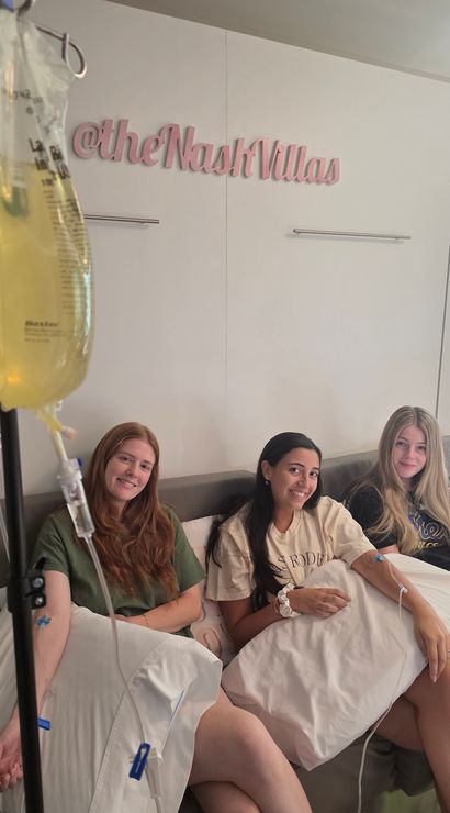 Three smiling women relax on a couch in a bright wellness lounge receiving IV vitamin drips, holding pillows while IV lines and yellow infusion bags hang in the foreground.