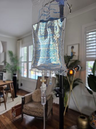 IV saline bag and drip chamber hanging from an IV pole in a bright, cozy living room with an armchair, potted plants, shuttered windows and hardwood floors — at-home IV setup in a residential interior.