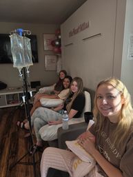 Group of four women relaxing in a cozy wellness lounge receiving IV drip therapy, seated with pillows, water bottle and smiles