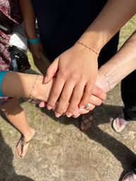 Three hands stacked in a sunny outdoor setting, delicate chain bracelets and a turquoise ring visible, casual sandals and grass in the background — a playful friendship gesture.