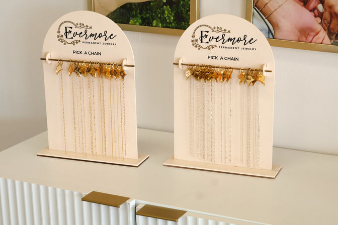 Two cream display stands on a white counter showcasing delicate gold and silver necklace chains with tiny star charms in a minimalist boutique setting