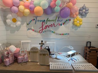Pastel balloon garland and daisy-shaped balloons frame a banner above a table filled with pink favor bags, jewelry display cards, a manicure lamp with tools and trays of rings — playful bridal shower decor.