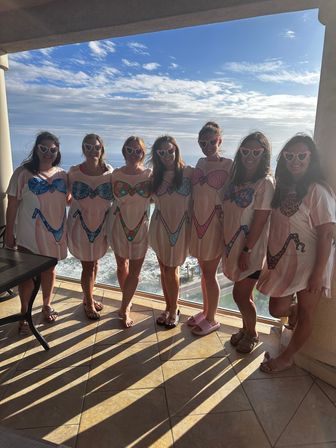 Group of seven women wearing matching bikini-print cover-up shirts and heart-shaped sunglasses posing on an oceanfront balcony overlooking sunlit waves and a blue sky