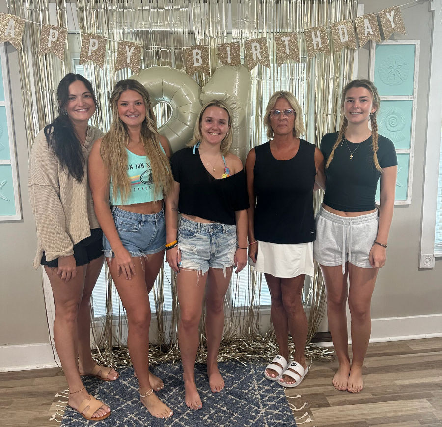 Five women posing indoors at a casual 21st birthday party with large gold '21' balloons, glitter 'Happy Birthday' banner and metallic fringe backdrop, wearing summer shorts and sandals