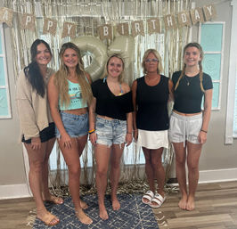 Five women posing indoors at a casual 21st birthday party with large gold '21' balloons, glitter 'Happy Birthday' banner and metallic fringe backdrop, wearing summer shorts and sandals