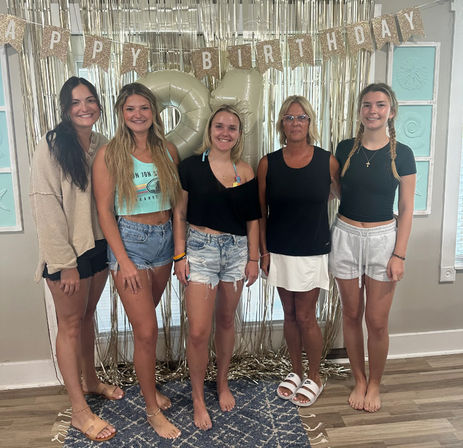 Five women posing indoors at a casual 21st birthday party with large gold '21' balloons, glitter 'Happy Birthday' banner and metallic fringe backdrop, wearing summer shorts and sandals