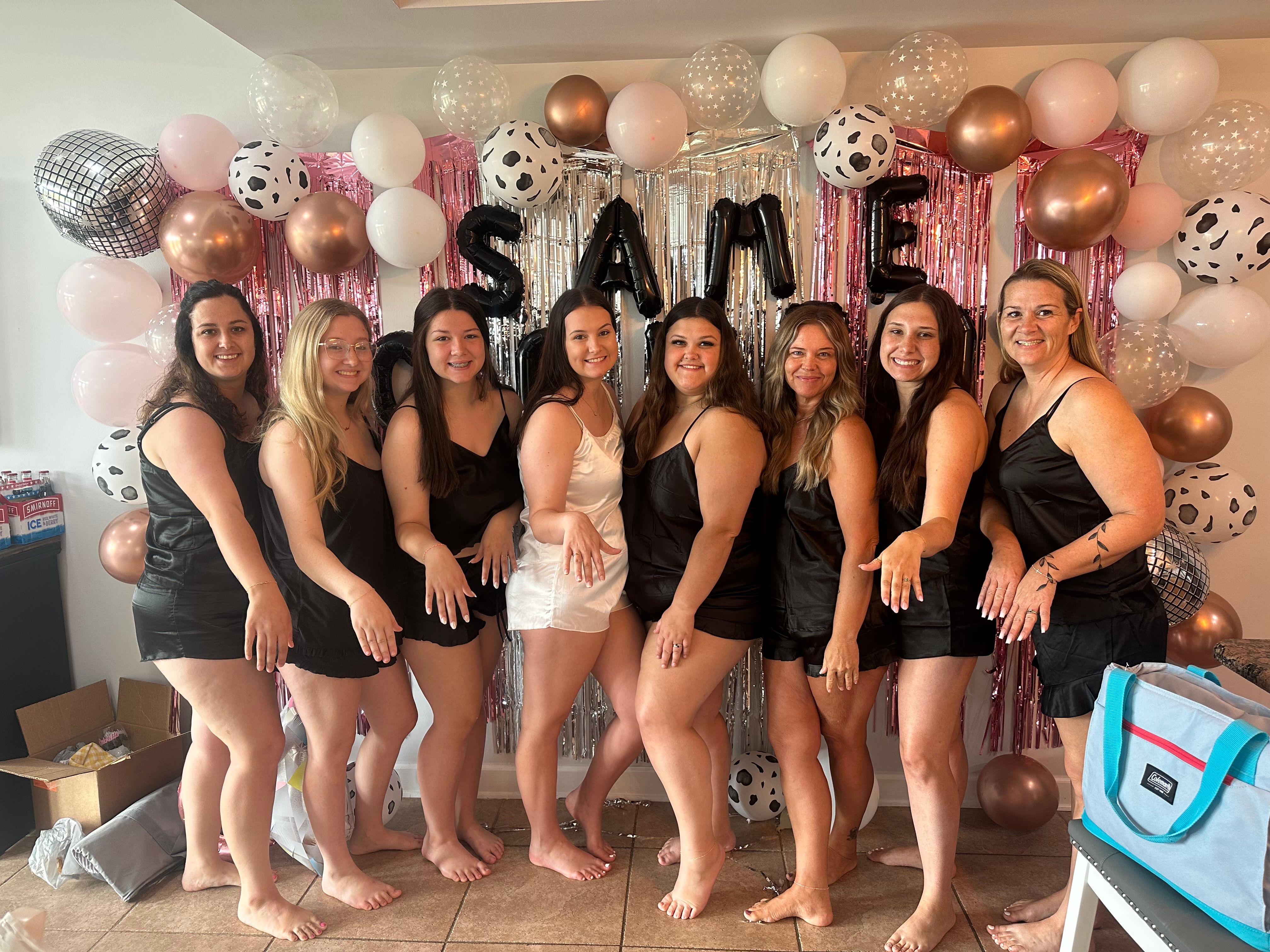 Bridal party: bride-to-be in a white slip dress and eight bridesmaids in matching black slip dresses posing barefoot and showing rings in front of a pink, silver and rose-gold balloon and fringe backdrop with metallic letter balloons for an indoor bachelorette celebration.