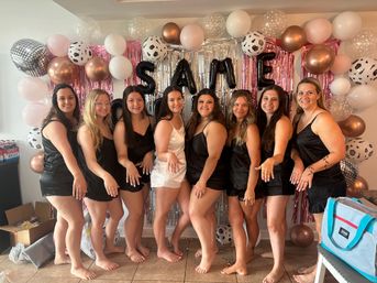 Bridal party: bride-to-be in a white slip dress and eight bridesmaids in matching black slip dresses posing barefoot and showing rings in front of a pink, silver and rose-gold balloon and fringe backdrop with metallic letter balloons for an indoor bachelorette celebration.