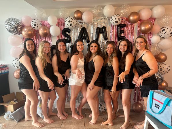 Bridal party: bride-to-be in a white slip dress and eight bridesmaids in matching black slip dresses posing barefoot and showing rings in front of a pink, silver and rose-gold balloon and fringe backdrop with metallic letter balloons for an indoor bachelorette celebration.