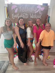 Five smiling friends in casual summer outfits show off rings in front of a colorful ribbon backdrop and a handwritten “Bachelorette Weekend” sign at an indoor celebration.