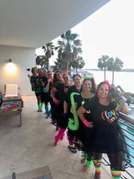 Group of women in neon 1980s-themed outfits and tutus posing on a beachfront resort balcony with palm trees and ocean view, holding inflatable saxophone, boombox and wearing bright leg warmers.